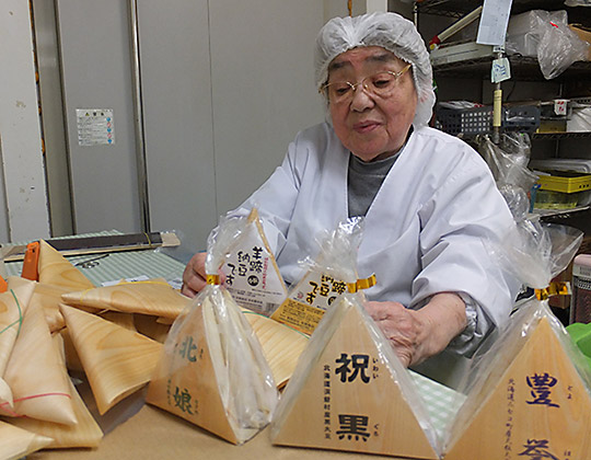 公式会社案内の作業風景3。経木包みの納豆を包装する工程を写す。